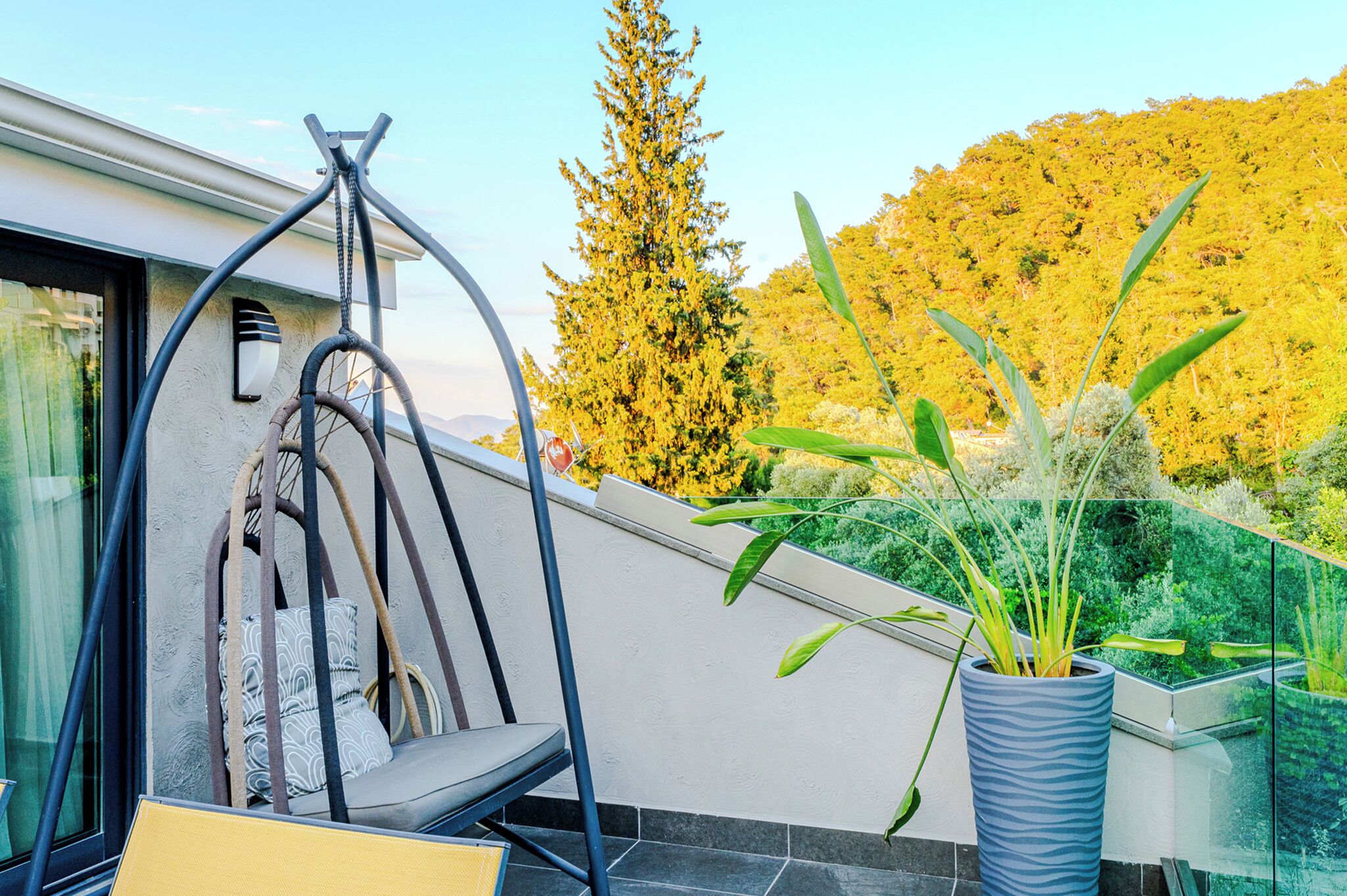 A modern outdoor seating area with a swing chair, a potted plant, and a view of a forested hillside.