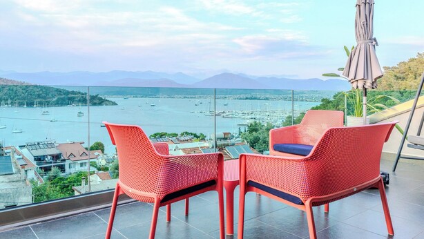 A balcony with red chairs and a table overlooking a marina with boats.