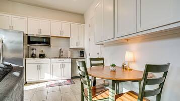 A modern kitchen with white cabinetry, a stainless steel refrigerator, a microwave, and a coffee maker. There is a dining table with chairs and a small potted plant on it.