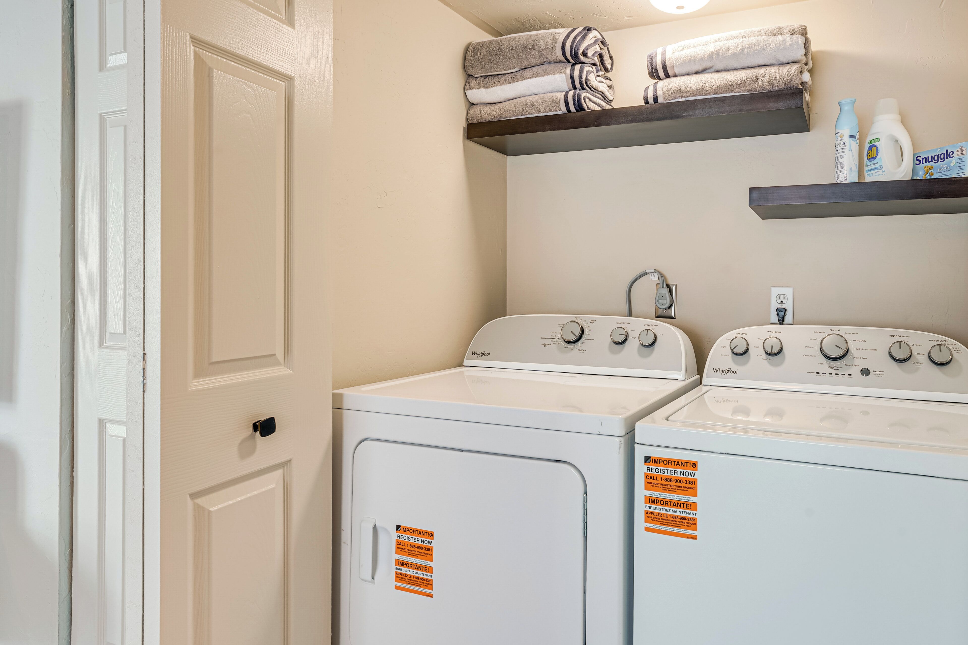 A laundry room with two white Whirlpool washers, a shelf with towels and cleaning products, and a door with a registration sticker.
