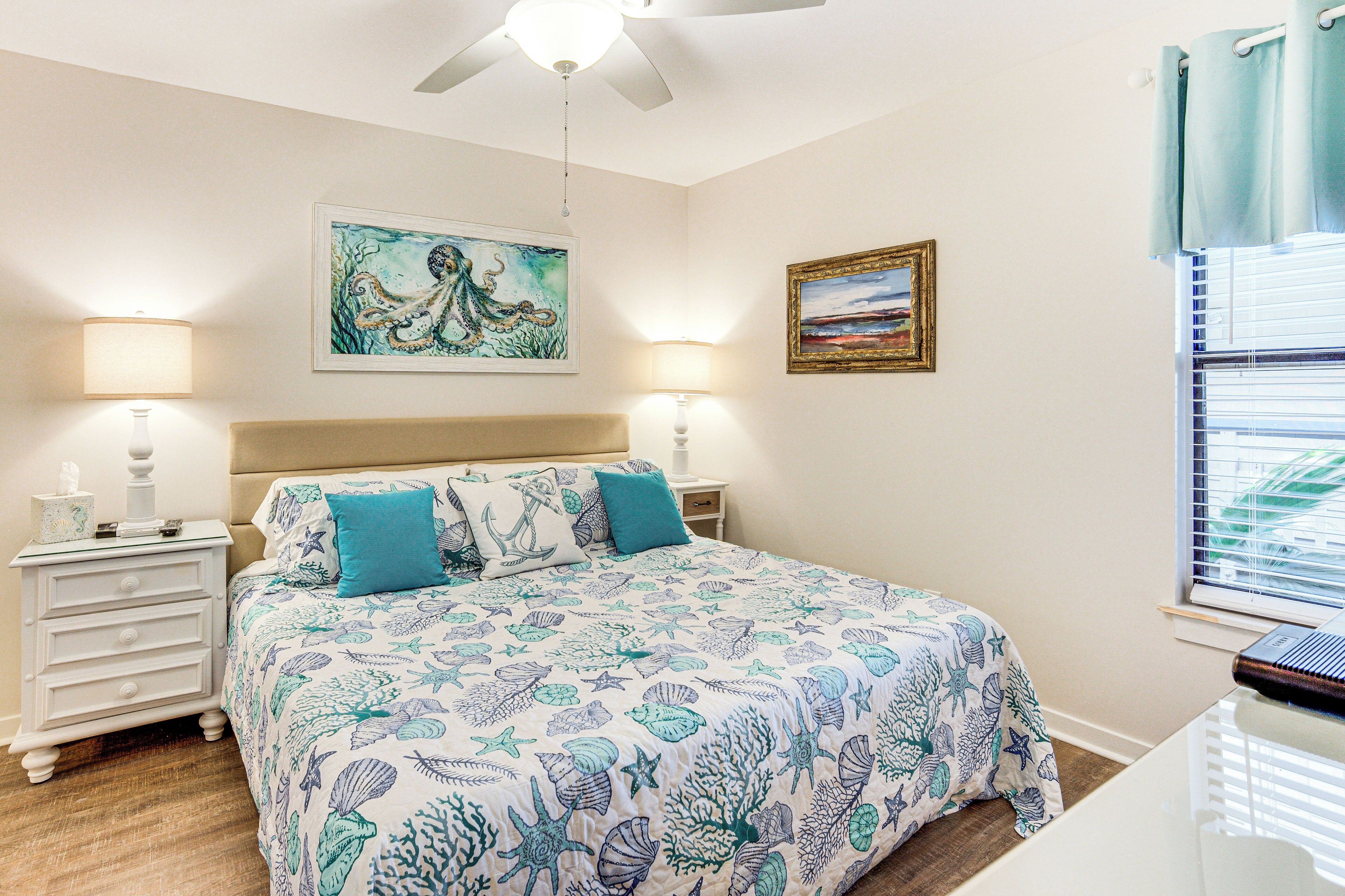 Une chambre à coucher comprenant un lit, deux tables de chevet, un ventilateur de plafond, une fenêtre avec des stores et des œuvres d’art murales.
