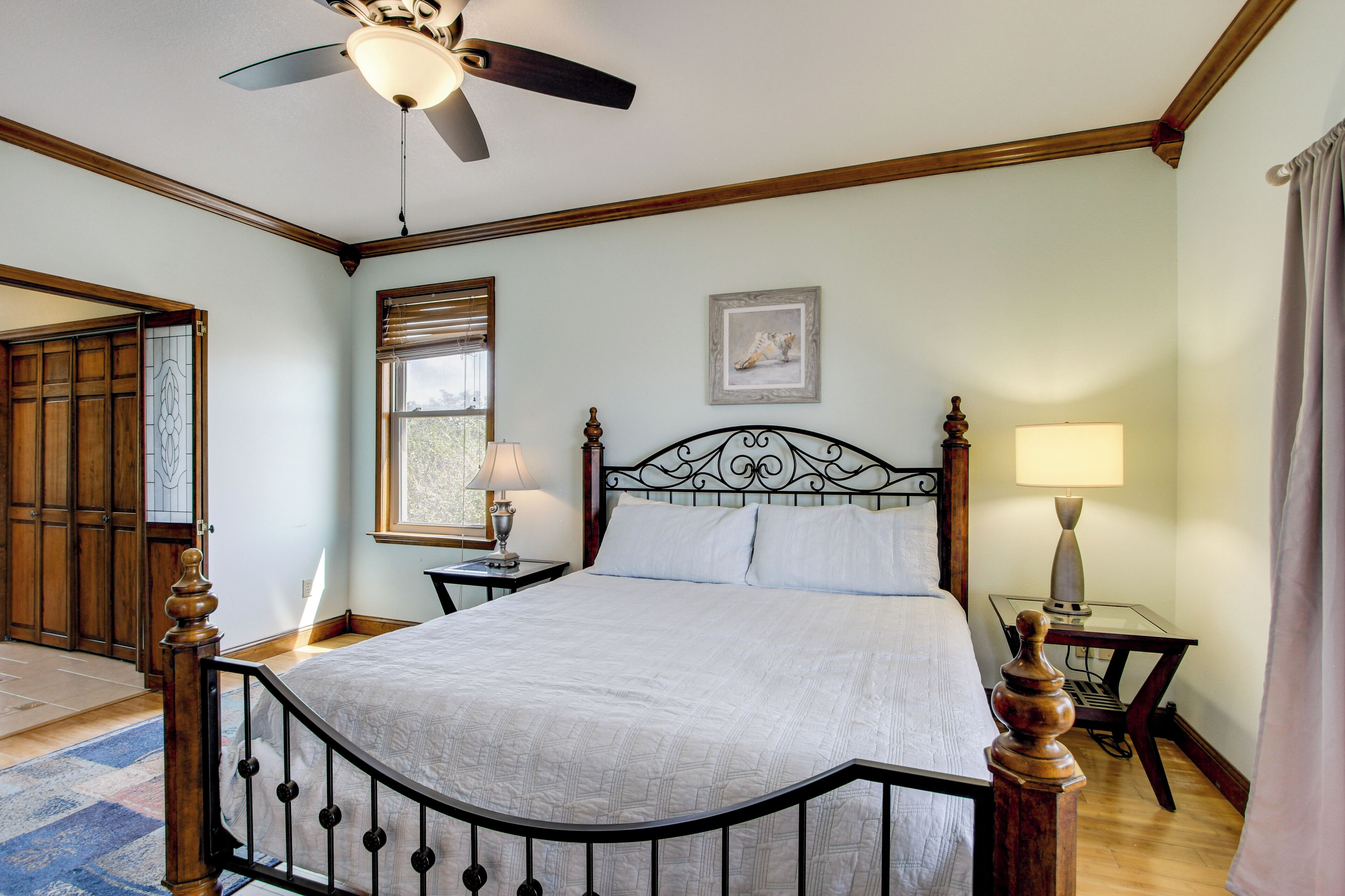 A bedroom with a bed, a ceiling fan, a window with blinds, a bedside table with a lamp, and a framed picture on the wall.