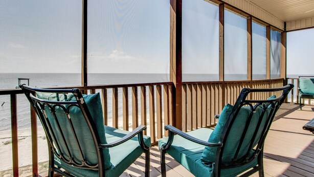 A covered outdoor seating area with two green cushioned chairs and a wooden railing overlooking the beach. - Private Beach & Views! Stilted Ocean Springs Home (Ocean Springs)