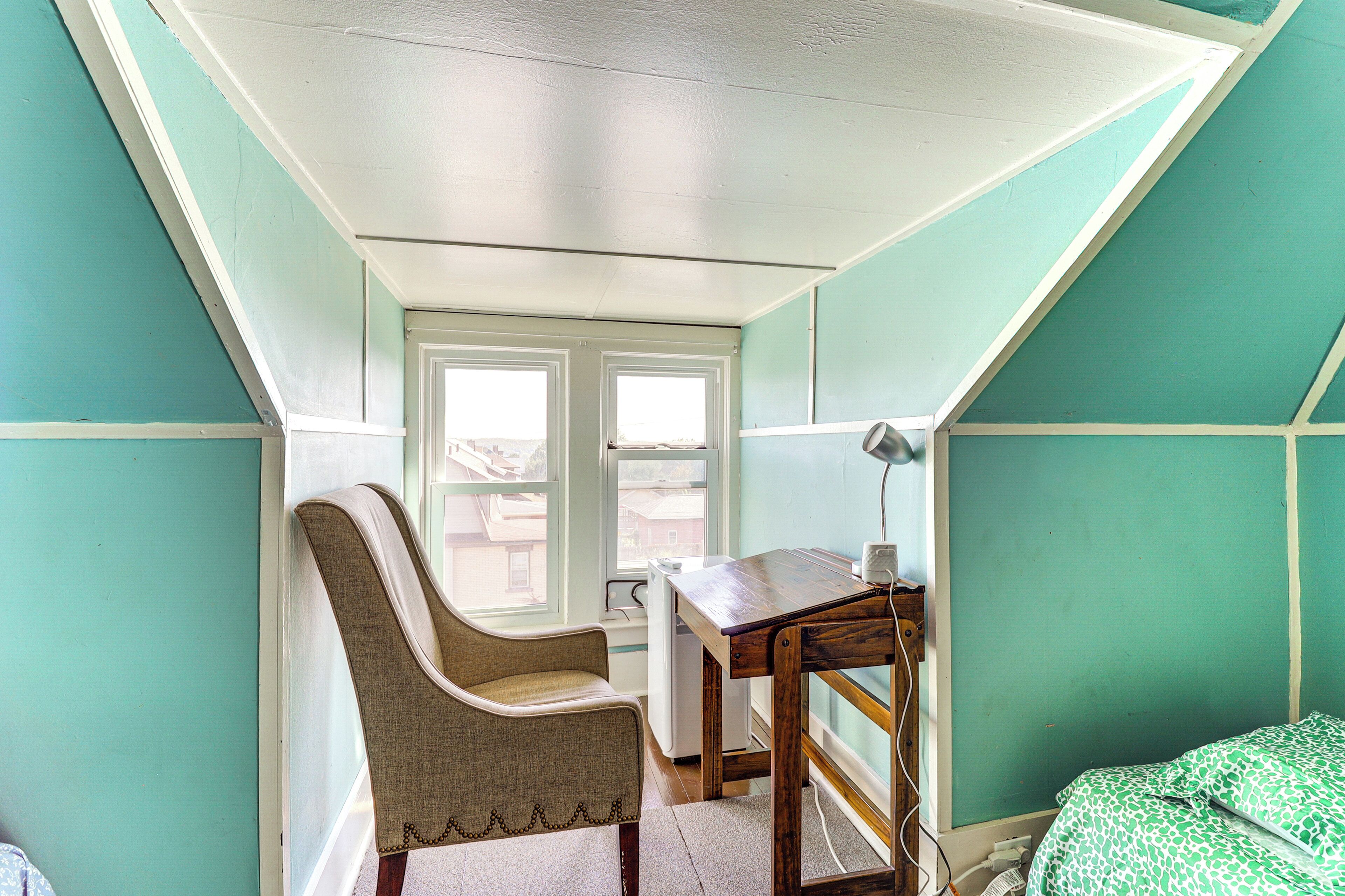 A room with teal walls, a wooden desk, a chair, a bed with patterned bedding, and a window with a view of buildings.