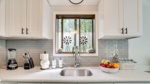 A kitchen with white cabinets, a stainless steel sink, a window with windmill decorations, and a bowl of fruit on the counter. - Yard + Game Room: Wooded Poconos Retreat (Tobyhanna)