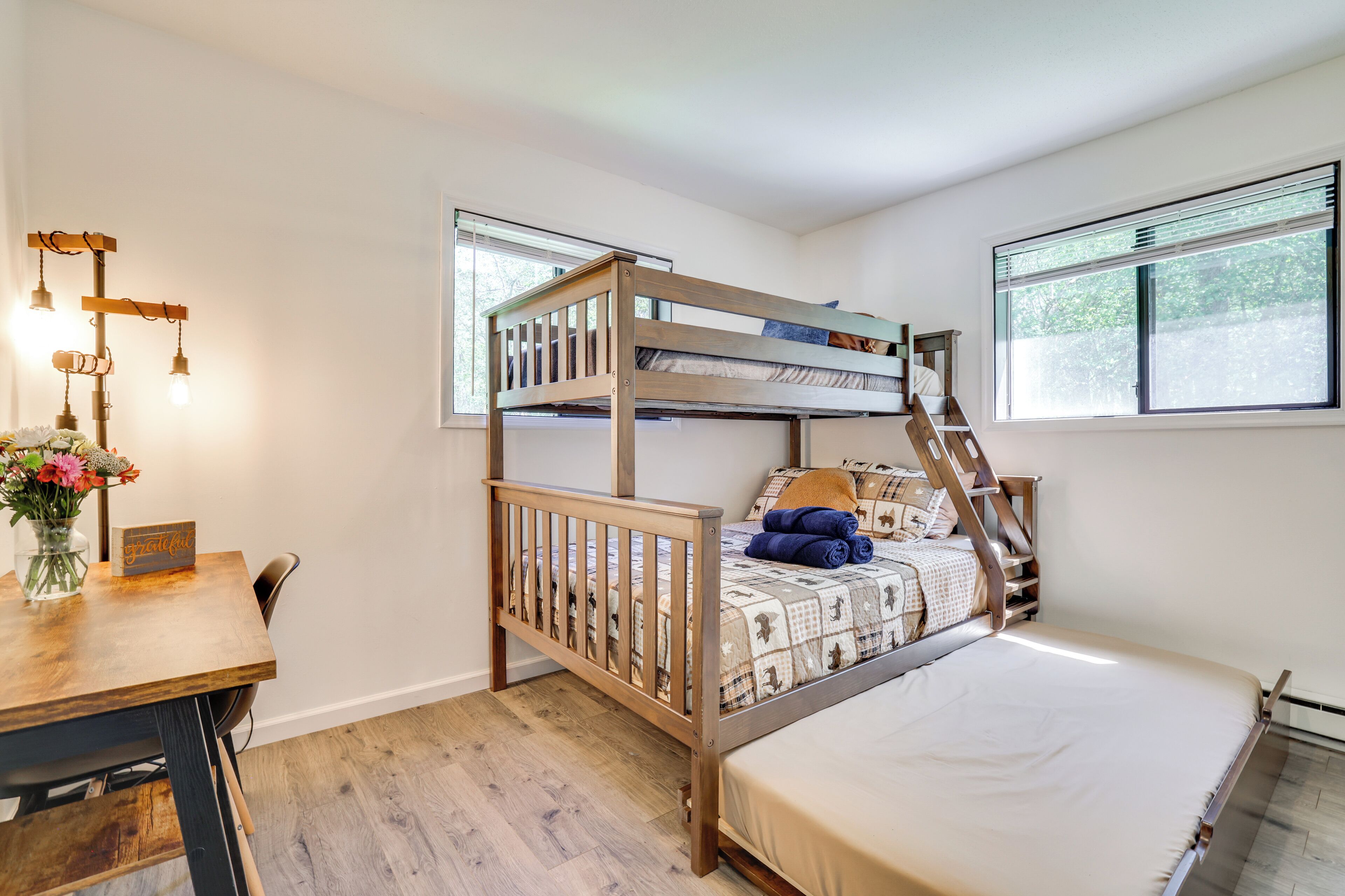 A bunk bed with a wooden frame, a desk with a vase of flowers, and a window with blinds.