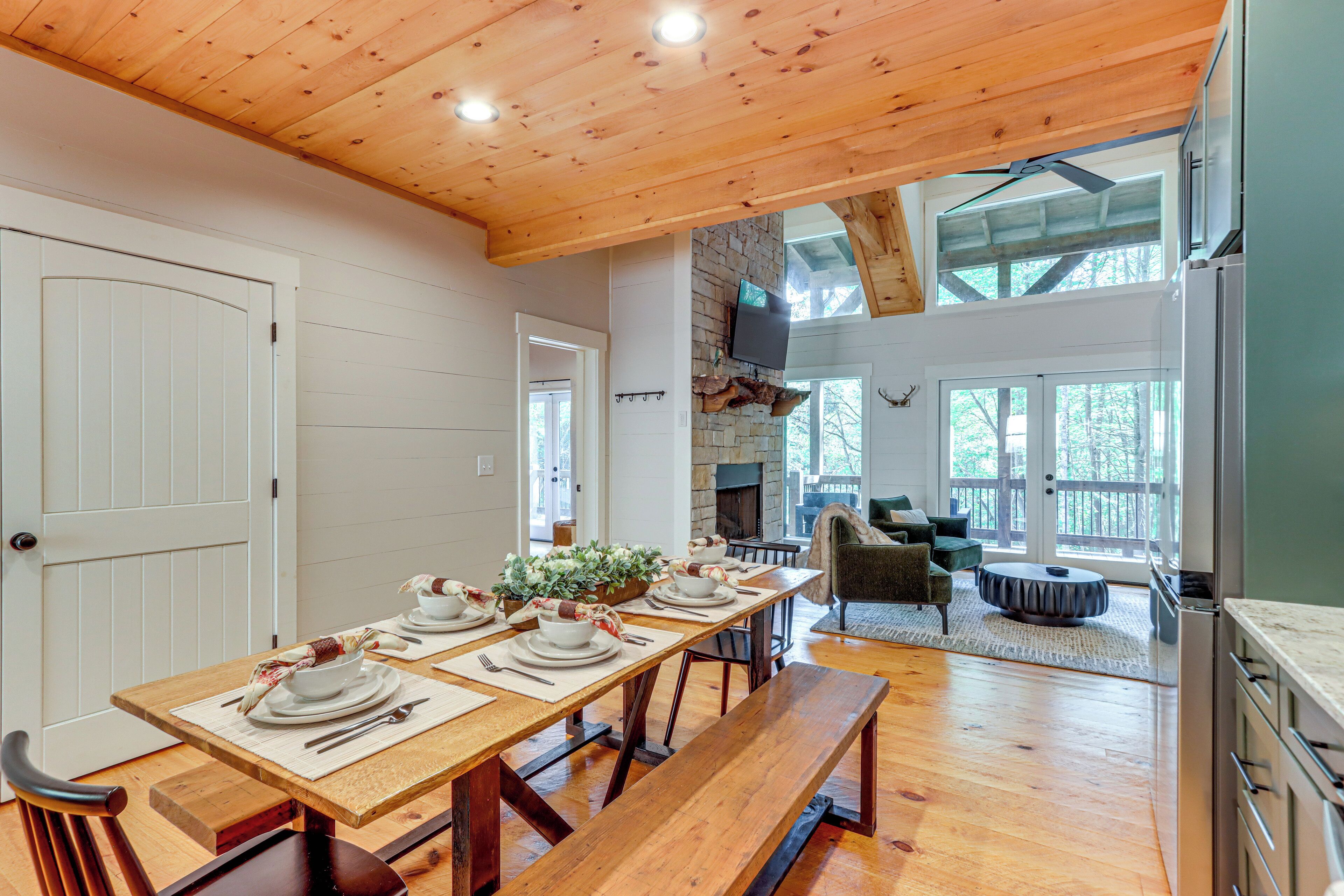 Un espace repas avec une longue table en bois dressée pour le repas, une cheminée en pierre et une vue sur l’extérieur grâce à de grandes fenêtres.