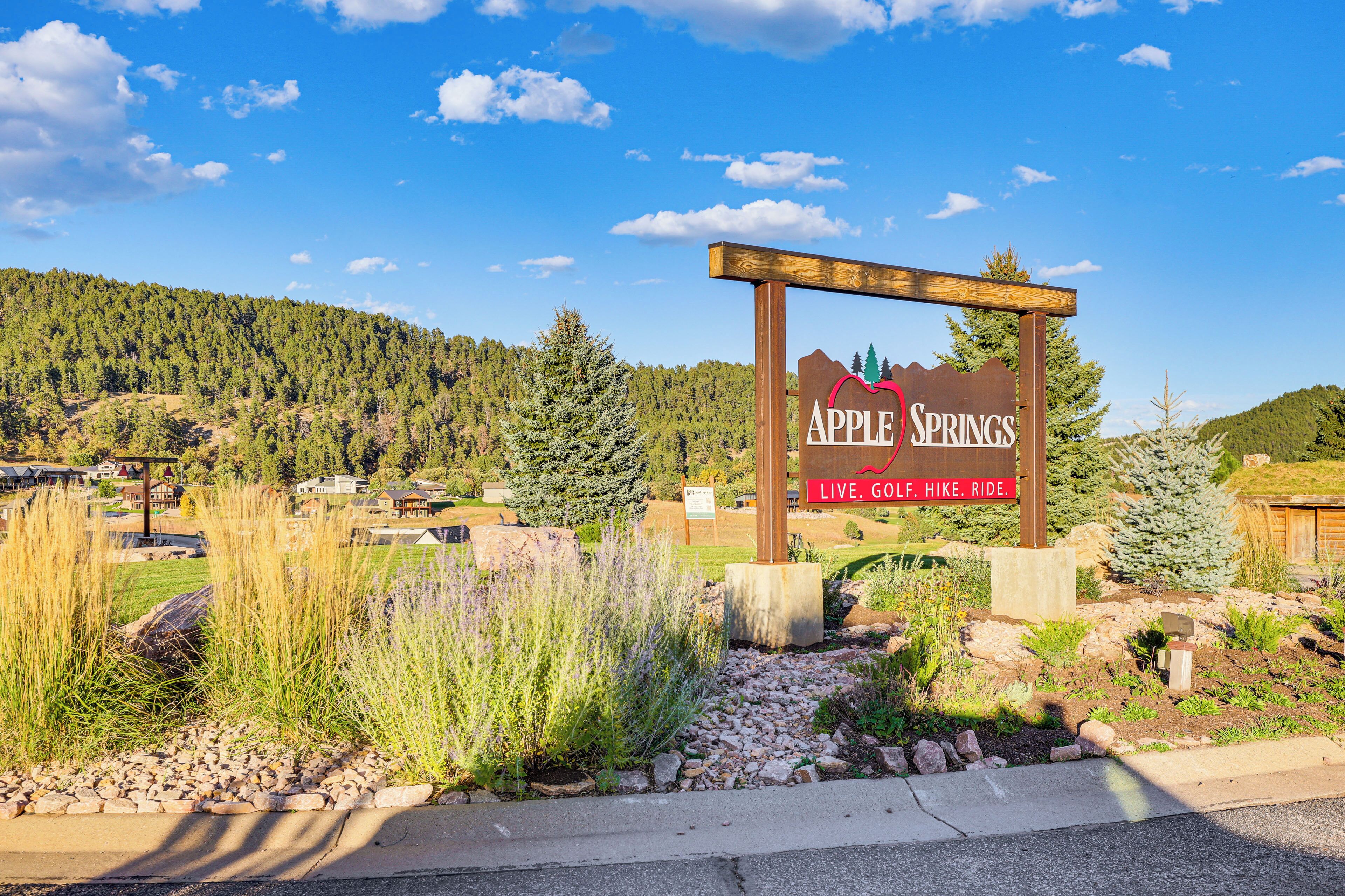 A wooden sign with the text "Apple Springs" and "Live. Go. Hike. Ride." in front of a mountainous landscape with houses in the background.