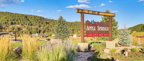 A wooden sign with the text "Apple Springs" and "Live. Go. Hike. Ride." in front of a mountainous landscape with houses in the background.