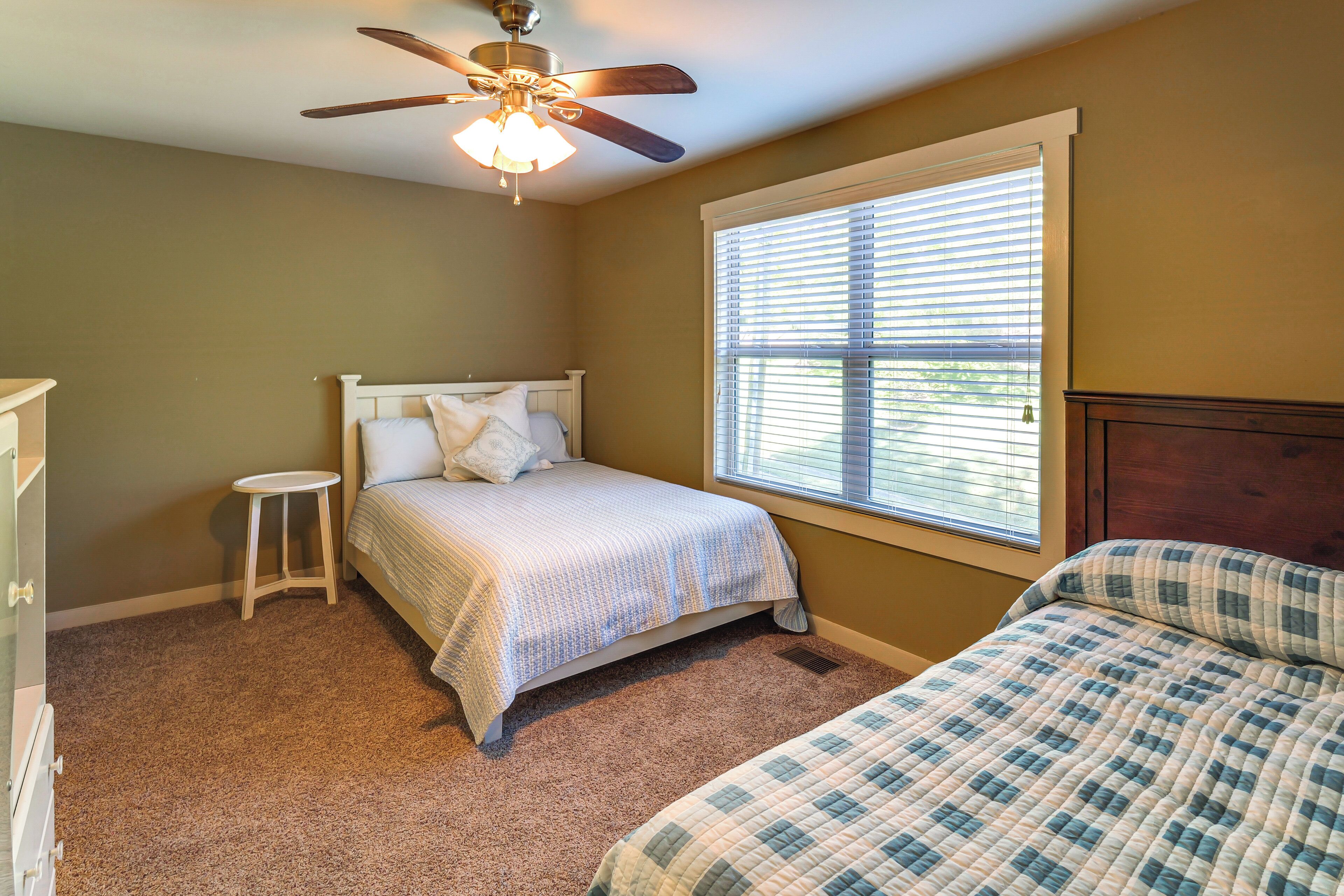 A bedroom with a bed, a small stool, a ceiling fan, and a window with blinds.