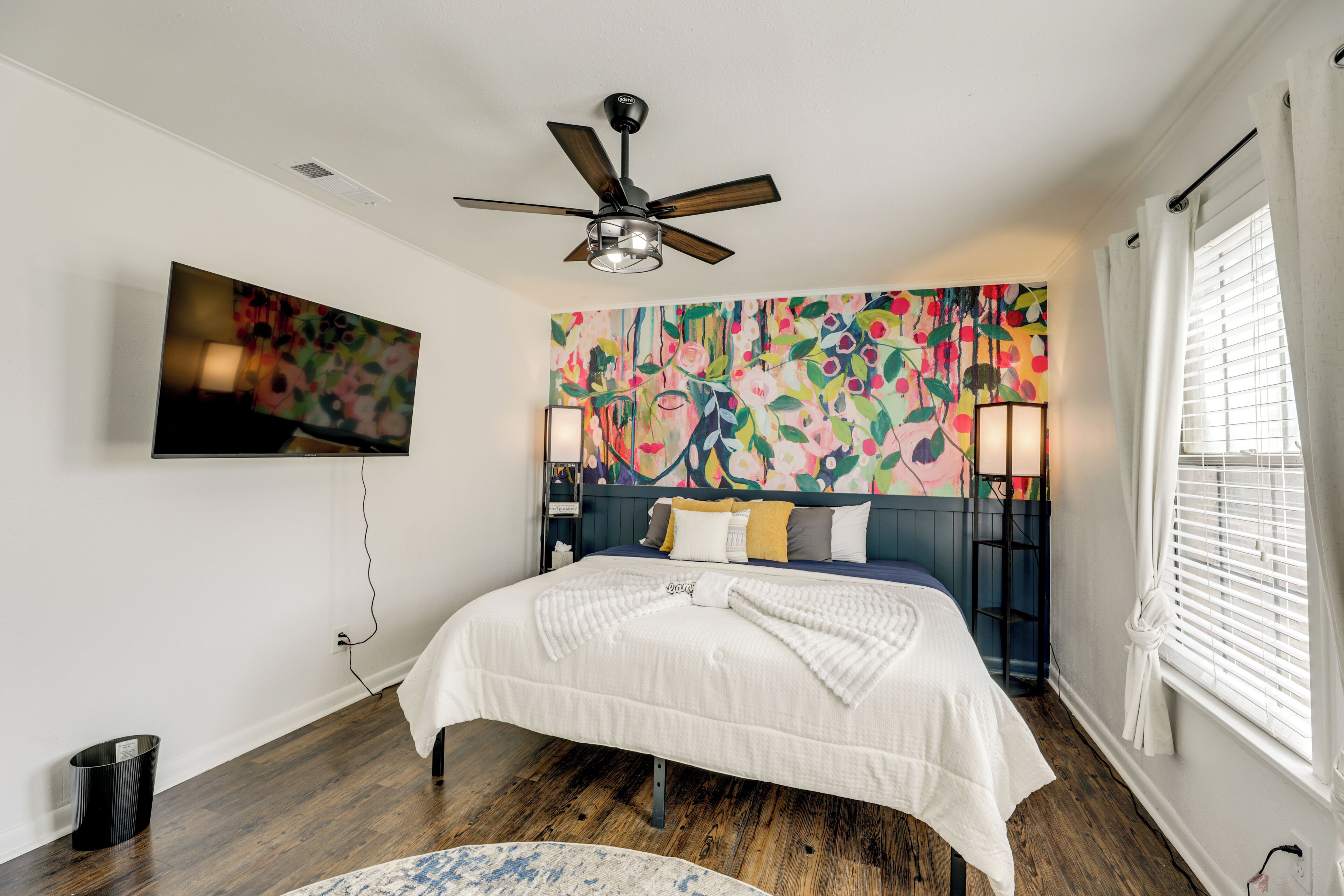Un dormitorio con cama, ventilador de techo, televisión montada en la pared y un mural floral colorido.