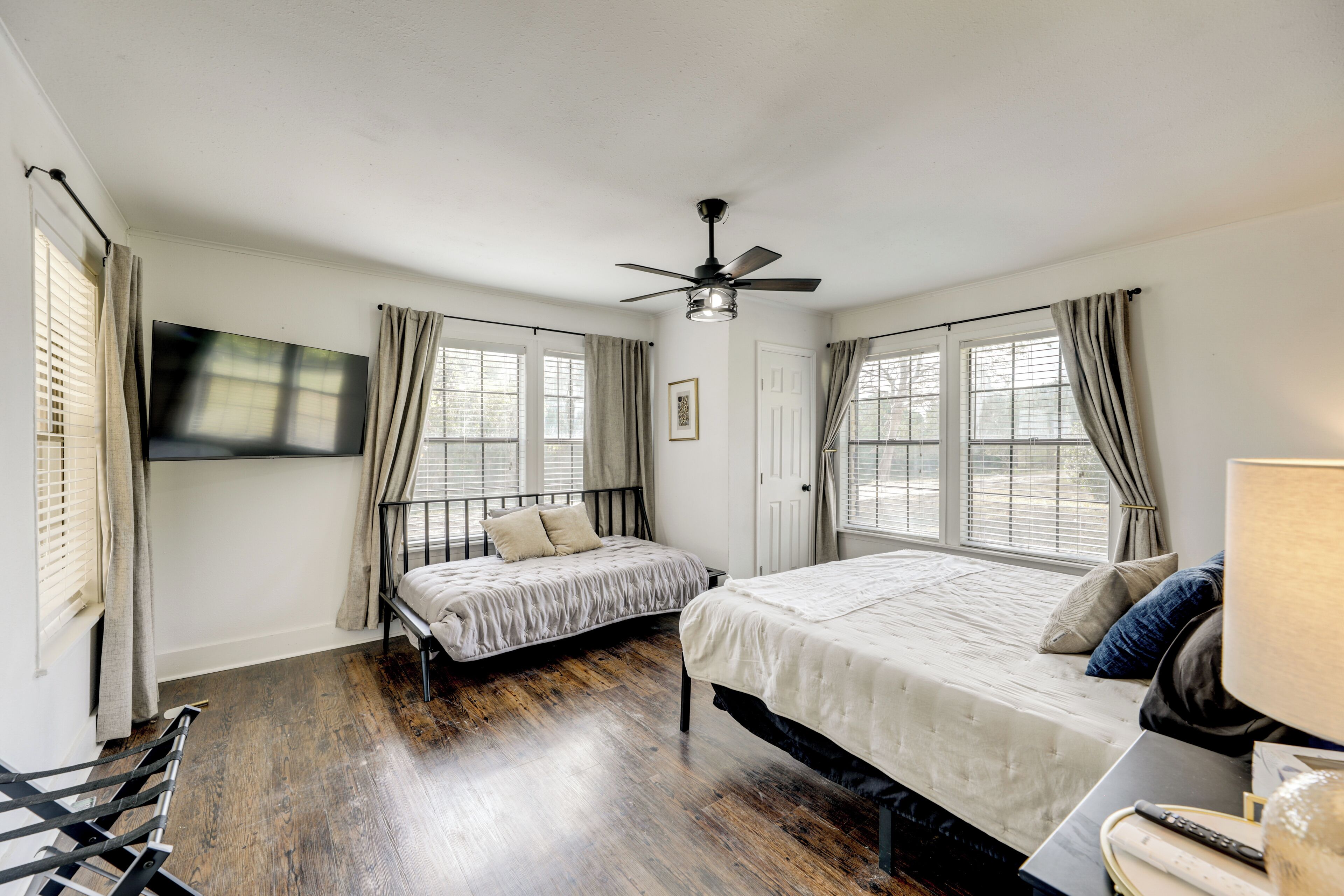 A bedroom with two beds, a ceiling fan, a TV mounted on the wall, and large windows with curtains.