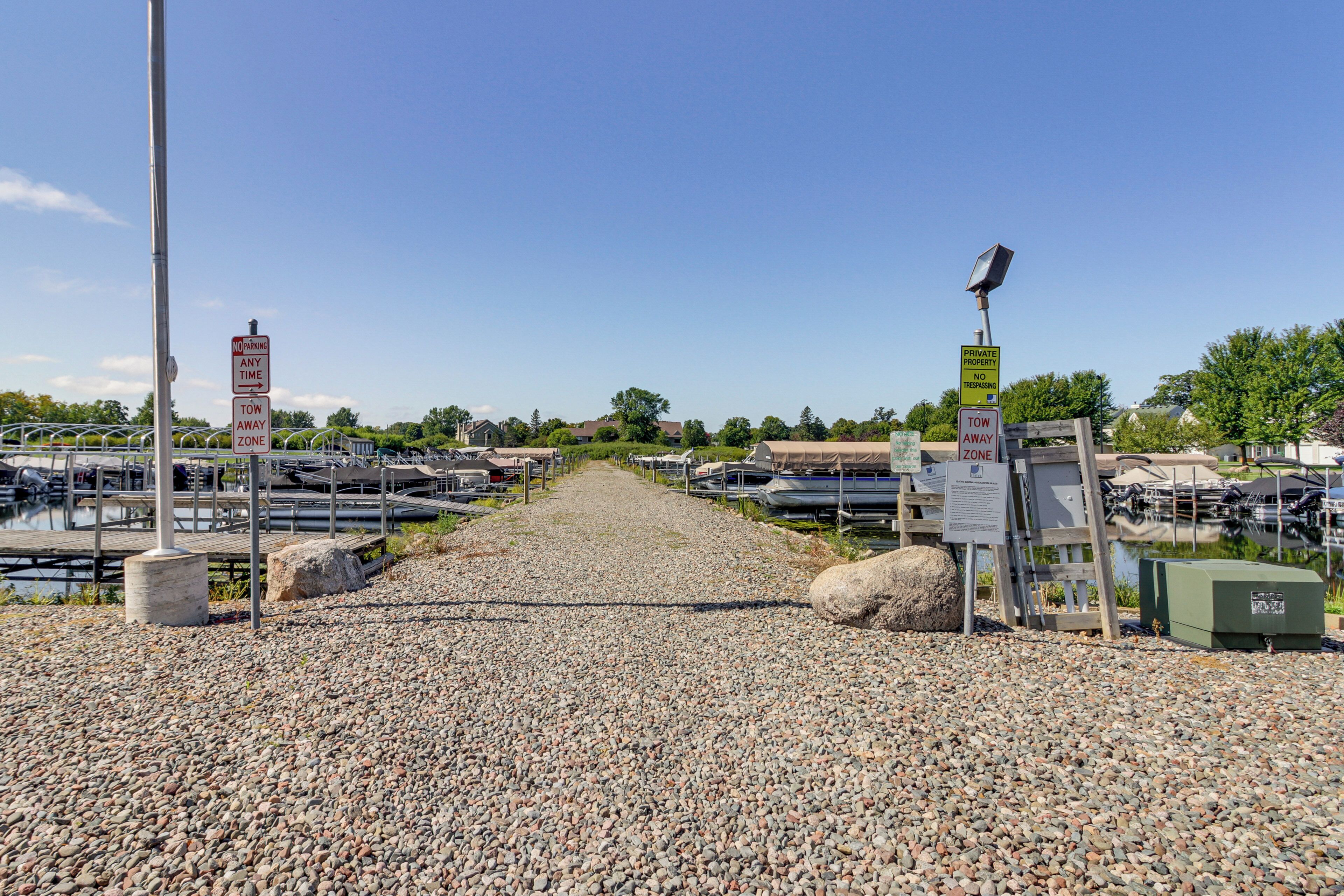 A gravel pathway leading to a marina with boats docked, surrounded by greenery and residential buildings.