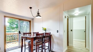 Un coin repas avec une table et des chaises en bois, un luminaire suspendu et une vue sur une terrasse en bois, accessible par une porte vitrée.
