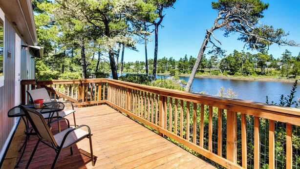 Une terrasse en bois, avec des chaises et une table, donnant sur un lac bordé d’arbres.