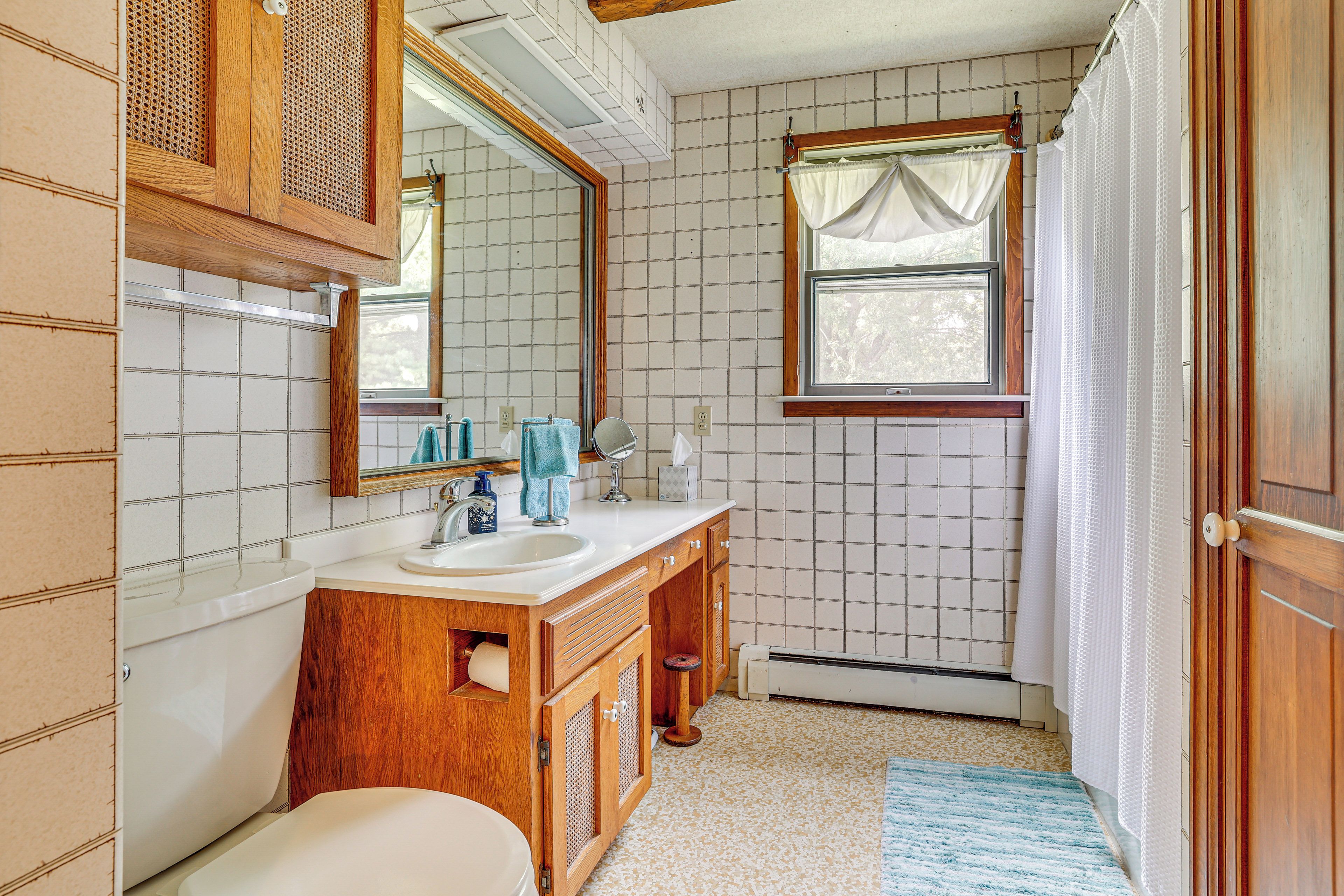A bathroom with a wooden vanity, a white toilet, a sink, a mirror, and a window with a curtain.