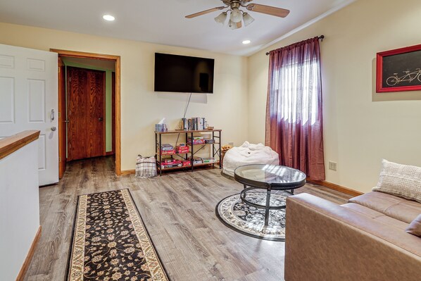 A living room with a flat-screen TV mounted on the wall, a round coffee table, a beige sofa, a wooden door, and a bookshelf with various items.