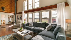 A spacious living room with a large sectional sofa, a coffee table, and a view of a field through large windows. - Mtn & Golf Course View: Oregon Outback Getaway! (Lakeview)