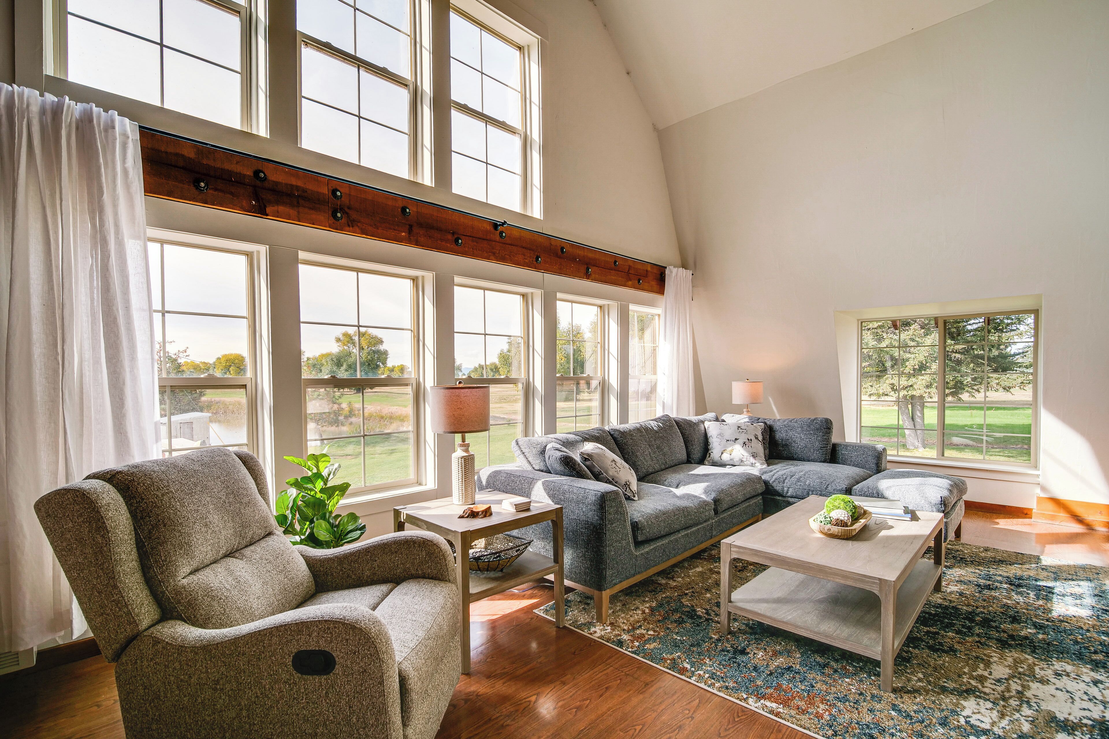 A spacious living room with a high ceiling, large windows, a grey sofa, a wooden coffee table, and a plant.