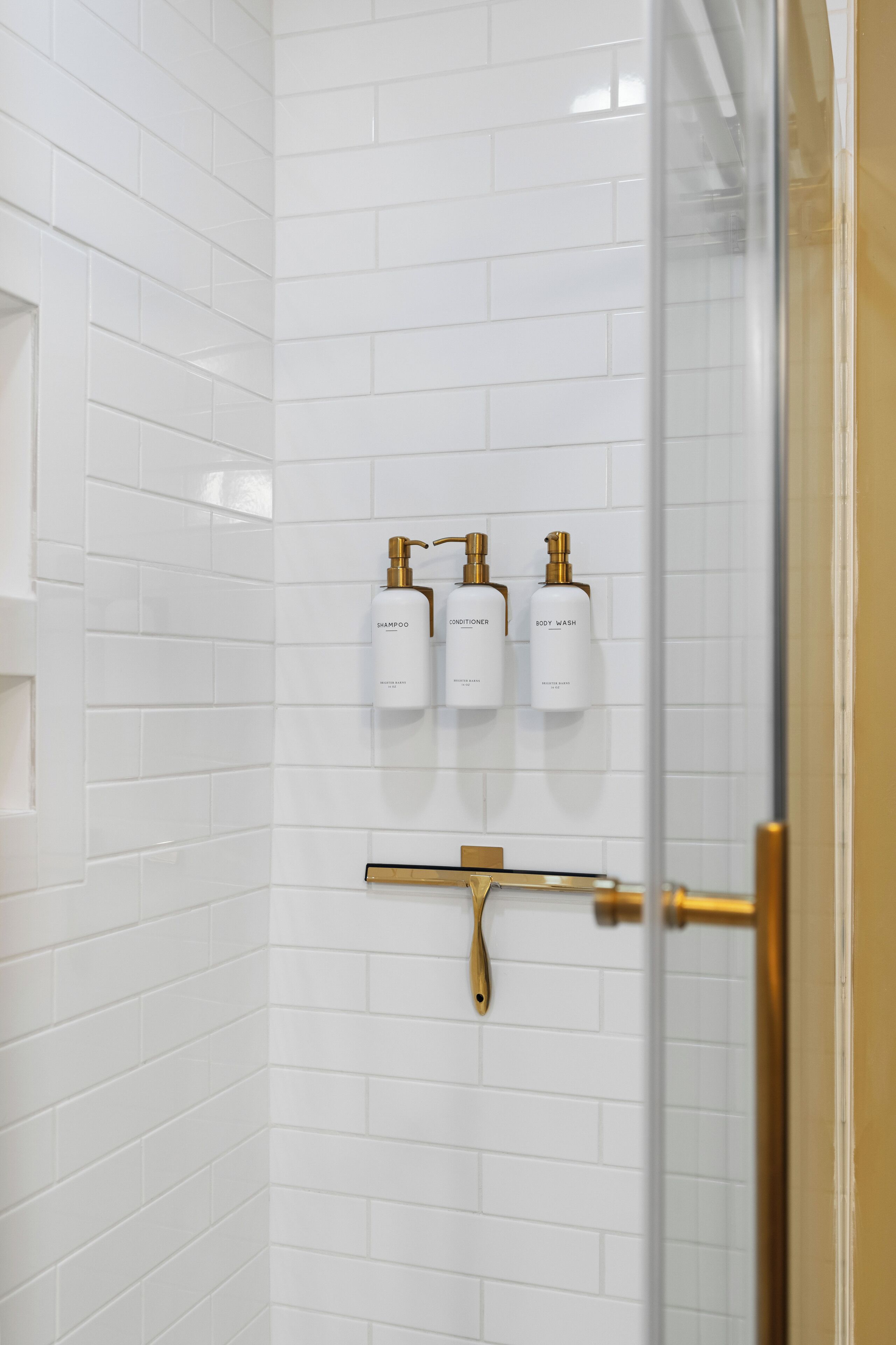 Ein Badezimmer mit weiß gefliesten Wänden, einer Duschkabine aus Glas und drei weißen Flaschen mit Shampoo, Conditioner und Duschgel.