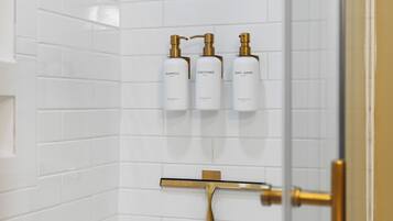 Ein Badezimmer mit weiß gefliesten Wänden, einer Duschkabine aus Glas und drei weißen Flaschen mit Shampoo, Conditioner und Duschgel.