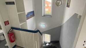 A staircase with a black mat at the bottom, a fire extinguisher on the left, and a small window with a view of a building outside. - EcoLodge2 (Oesterdeichstrich)