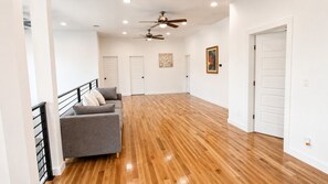 Un amplio espacio de sala con piso de madera, un sofá gris, un ventilador de techo y puertas blancas.