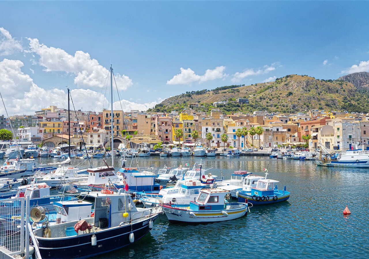 A marina with numerous boats docked, surrounded by colorful buildings and a hilly backdrop.