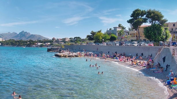Egy tiszta, türkizkék vízű strand, ahol az emberek úsznak és napoznak, a háttérben pedig épületek láthatók.