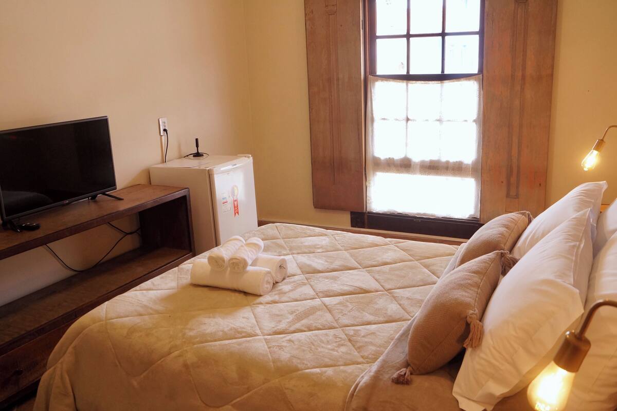 A hotel room with a bed, a television, a small refrigerator, and a window with curtains.