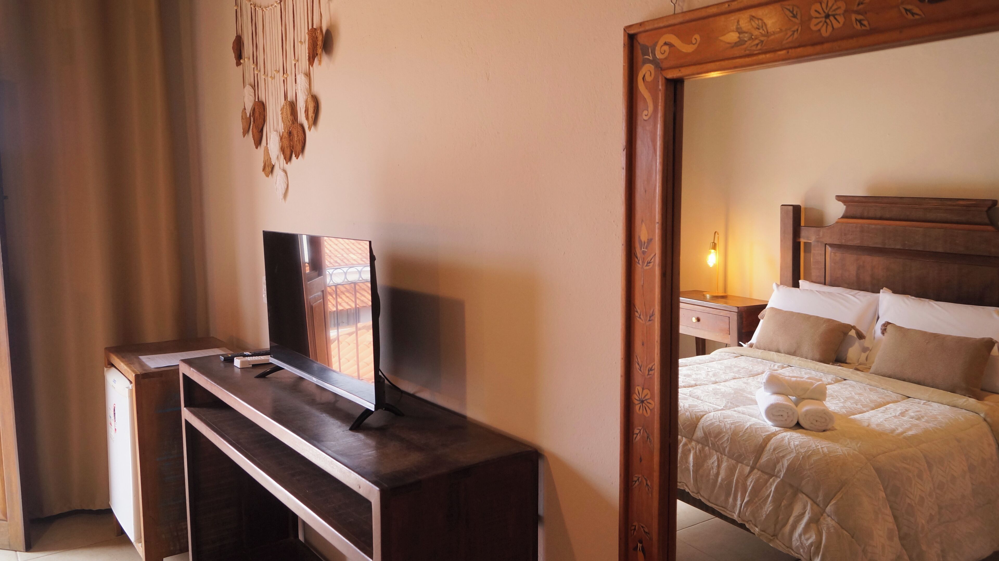 Habitación de hotel con cama, mesita de noche de madera con lámpara, televisor sobre un soporte de madera y espejo con marco decorativo.