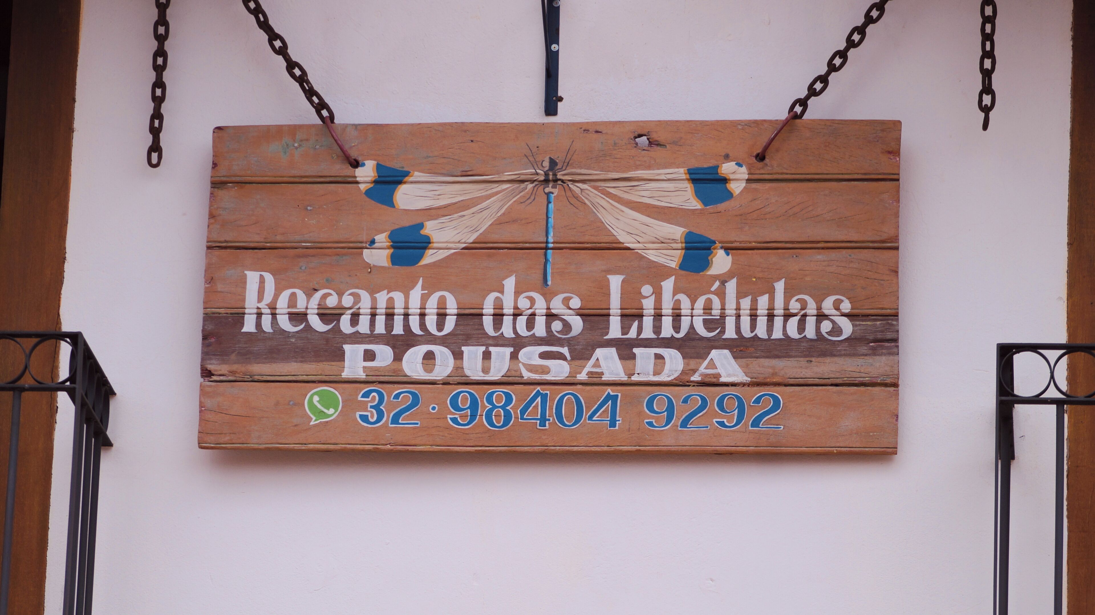A wooden sign with a dragonfly design and text reading "Recanto das Libélulas Pousada" along with a phone number.