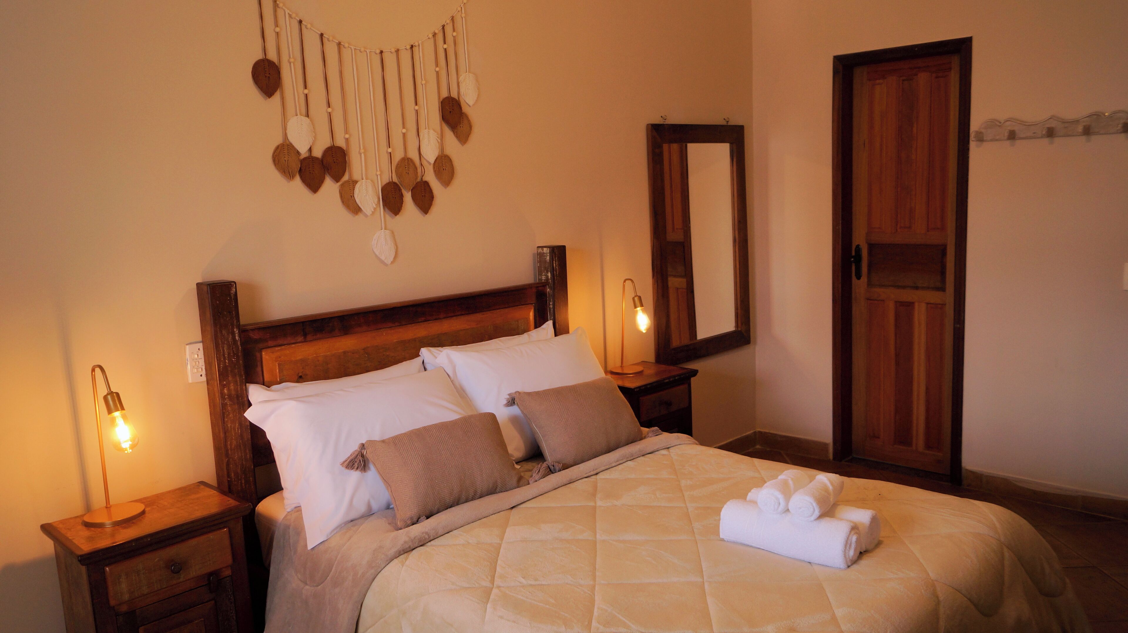 A neatly made bed with a quilted cover, two bedside lamps, a wooden headboard, a wall hanging of leaves, and a mirror.