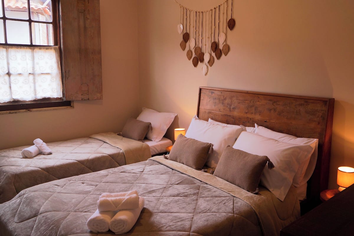 A room with two beds, each with a quilted bedspread and pillows, a wooden headboard, a wall hanging, and a window with a curtain.