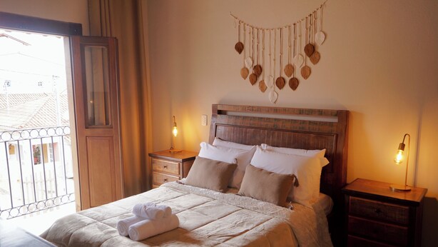 A neatly made bed with white linens and a decorative wall hanging above it. There are two bedside tables with lamps, and a window with curtains to the left. - Recanto das Libelulas pousada (Tiradentes)