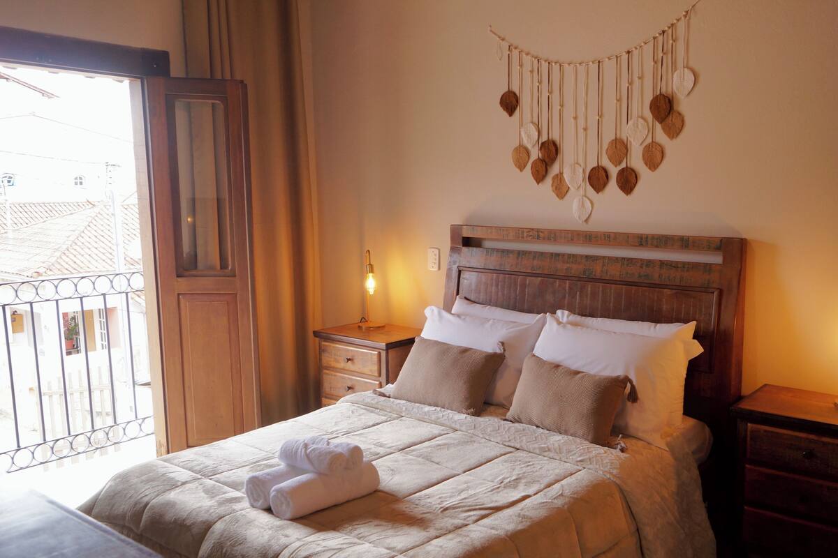 A neatly made bed with white linens and a decorative wall hanging above it. There are two bedside tables with lamps, and a window with curtains to the left.