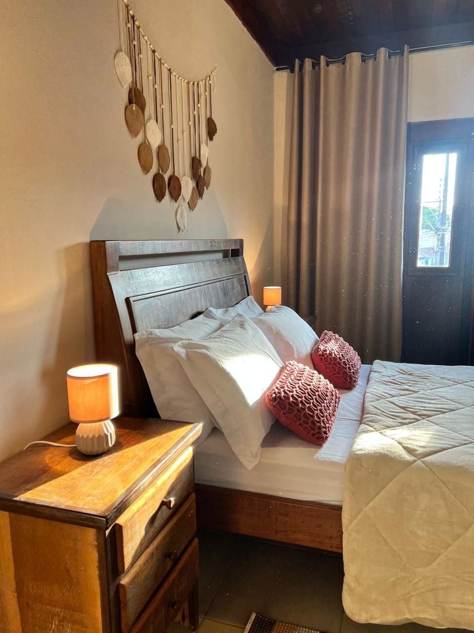 Cama bem arrumada com lençóis brancos e duas almofadas cor-de-rosa, mesa de cabeceira de madeira com abajur e um painel de parede com folhas penduradas.