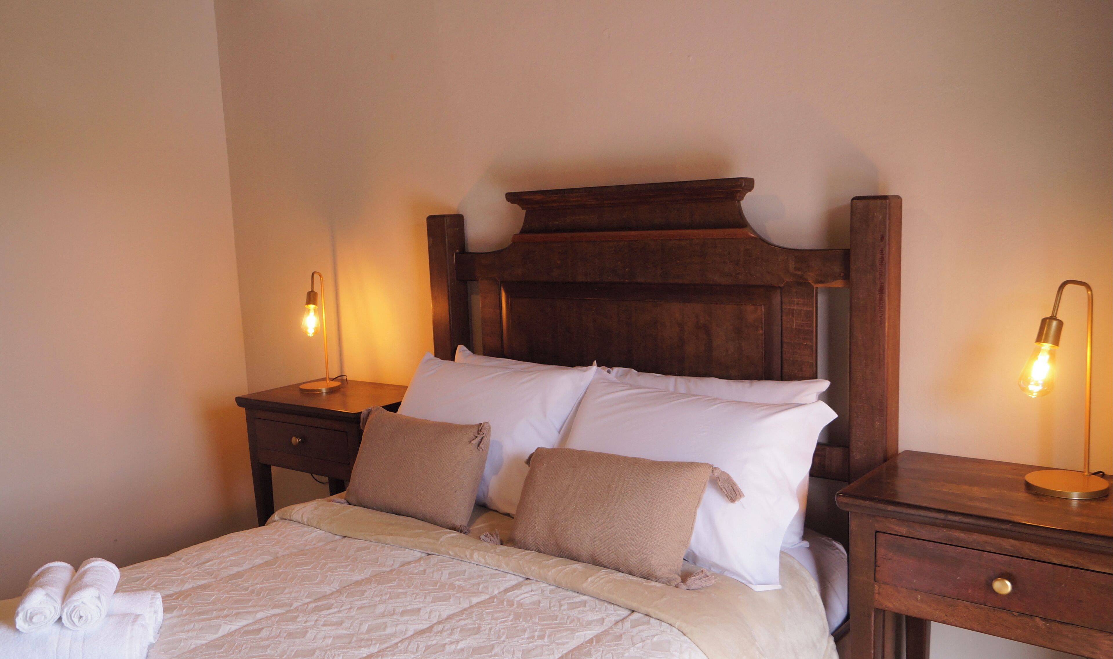 A neatly made bed with white and beige linens, two bedside tables with lamps, and a wooden headboard.