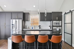 A modern kitchen with dark cabinets, stainless steel appliances, a central island with bar stools, and a window with blinds.