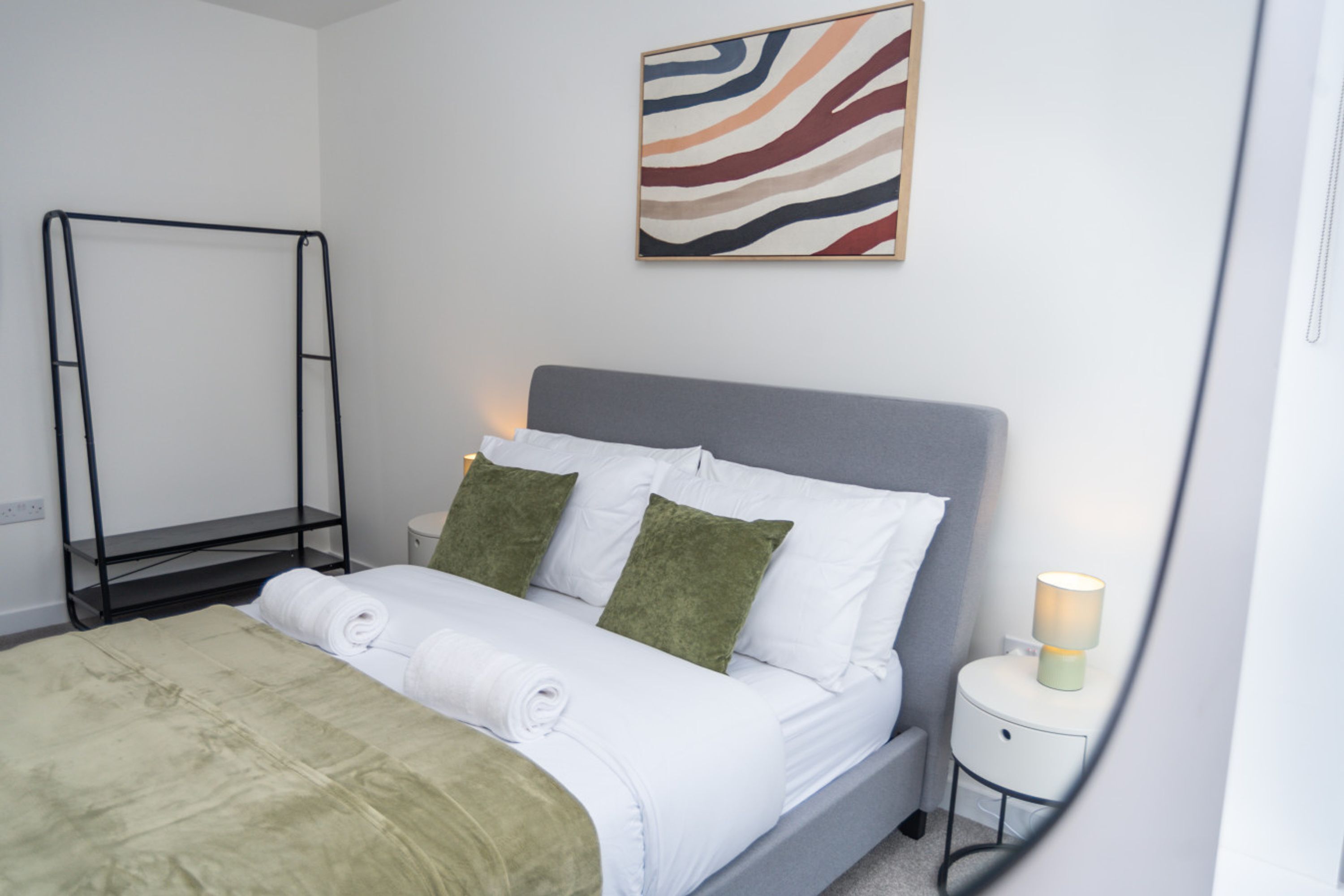 A neatly made bed with white linens and green pillows, a gray headboard, a bedside table with a lamp, and a wall-mounted abstract art piece.