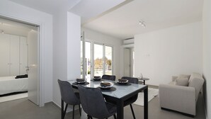 A modern dining area with a table set for six, surrounded by chairs. There is a sofa and a small table with a vase on the right side. - Perdika Residences (Aegina)