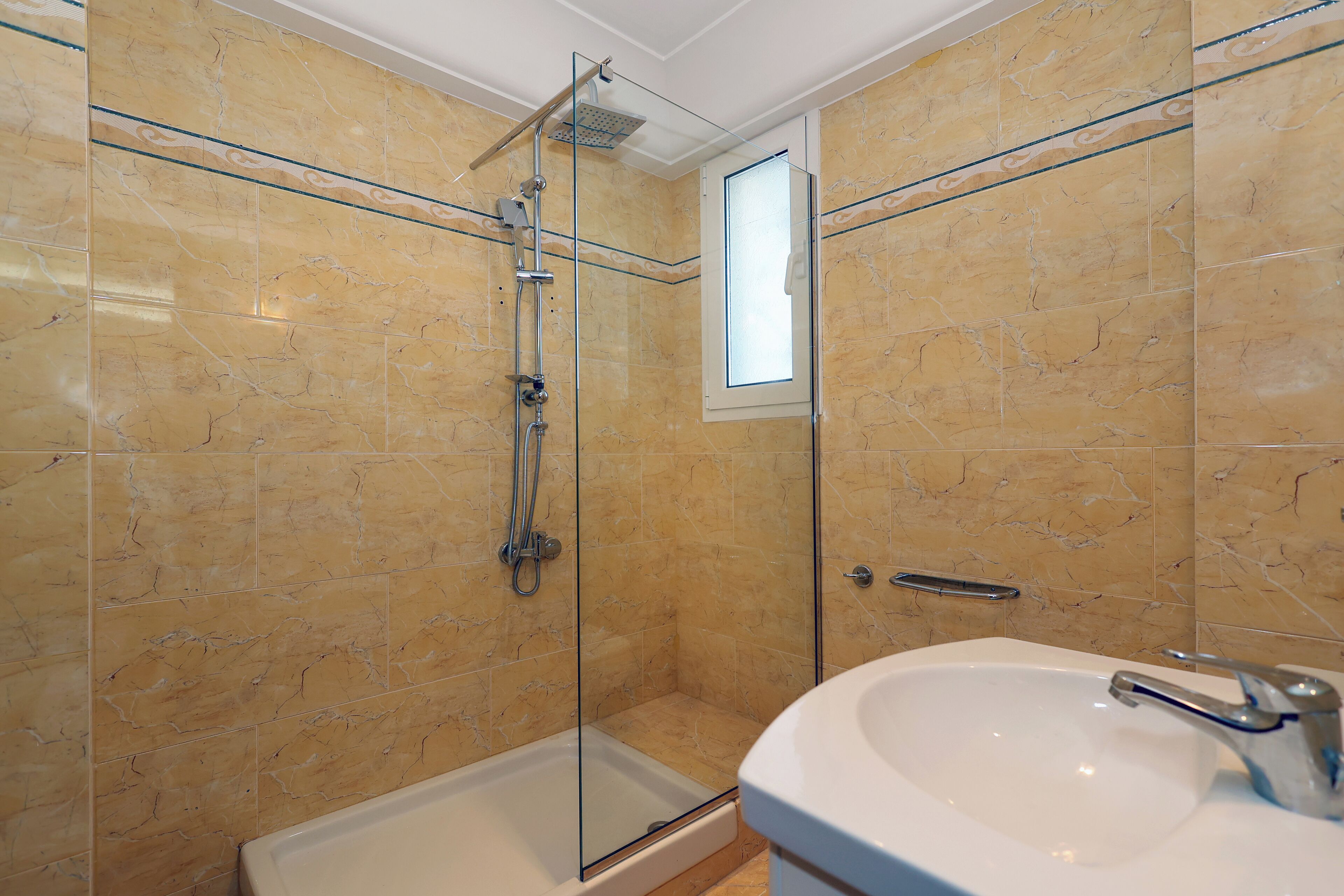 A bathroom with a glass-enclosed shower, a white sink, and beige marble tiles.
