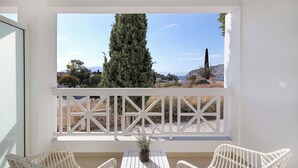 A balcony with white wicker chairs and a table, overlooking a landscape with trees and hills. - Perdika Residences (Aegina)