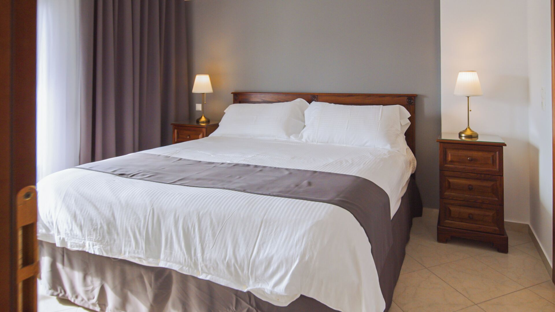 Una cama bien hecha con ropa de cama blanca y gris, dos mesitas de noche con lámparas y un cabecero de madera.