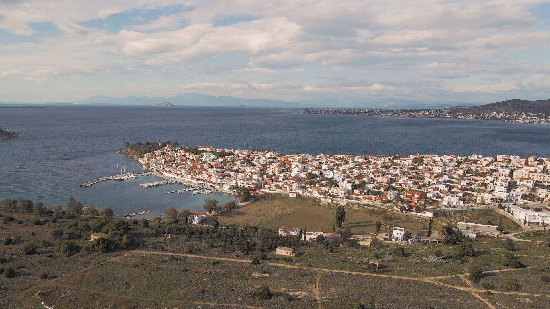 Vista aerea di una cittadina costiera con un porto turistico, edifici densi e colline circostanti.