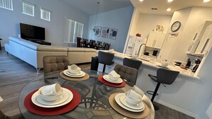 Uma sala de estar moderna com uma mesa de jantar de vidro posta para quatro pessoas, um sofá e uma televisão.