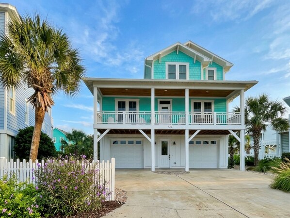 A two-story house with a balcony, a garage, and a white picket fence. - Seahorse by the Sea | Modern Comforts, Outdoor Living + Beach Access (Kure Beach)