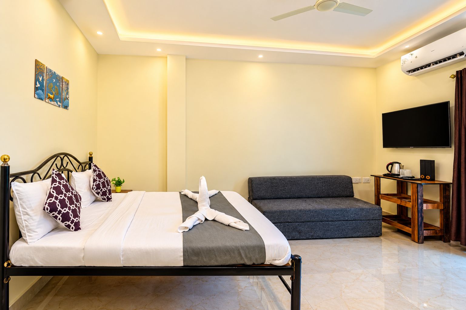 A hotel room with a bed, a sofa, a TV, and an air conditioning unit.
