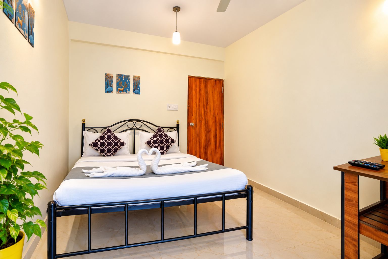 Habitación de hotel con cama, puerta de madera, mesita con una planta y pared con tres cuadros.
