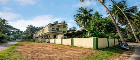 A residential building with a flat roof, a covered porch, and a fenced yard with grass and palm trees.